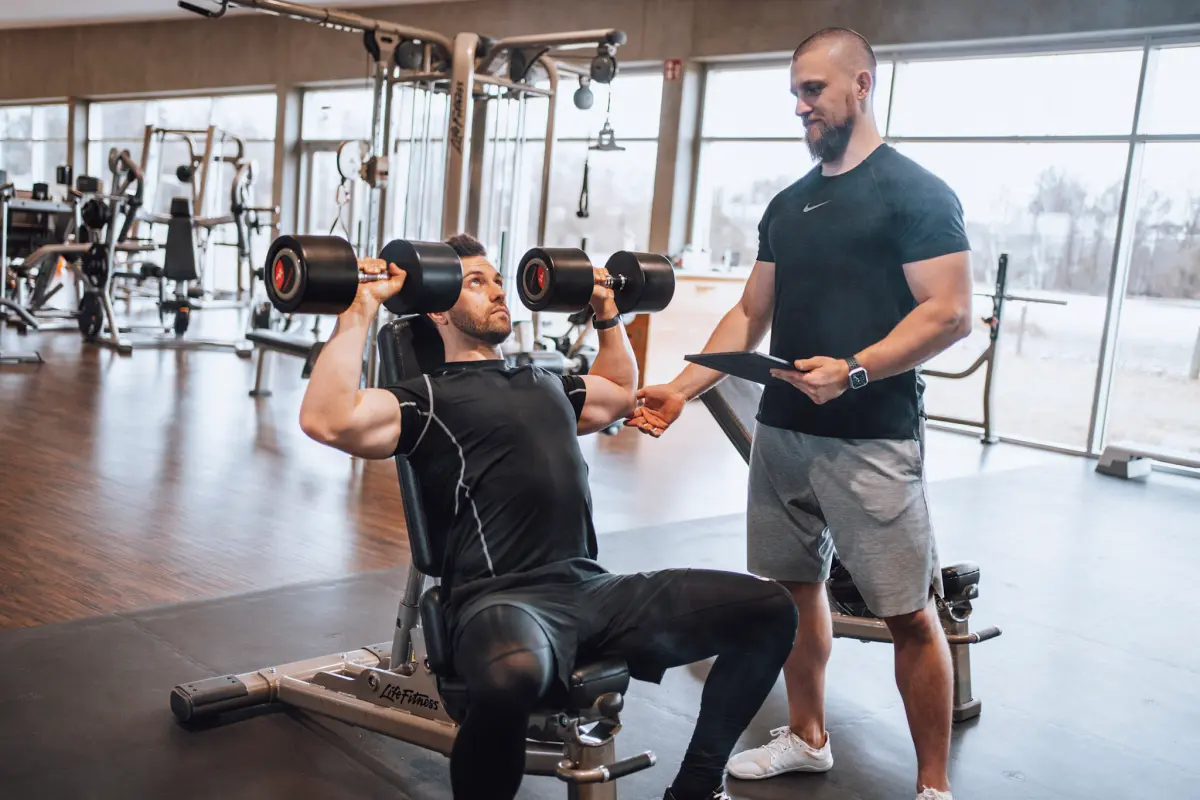 David Bachmeier mit einem seiner Klienten im Fitnesstudio, der gerade mit Gewichten auf der Bank sitzt und sein Training zum Muskelaufbau absolviert.
